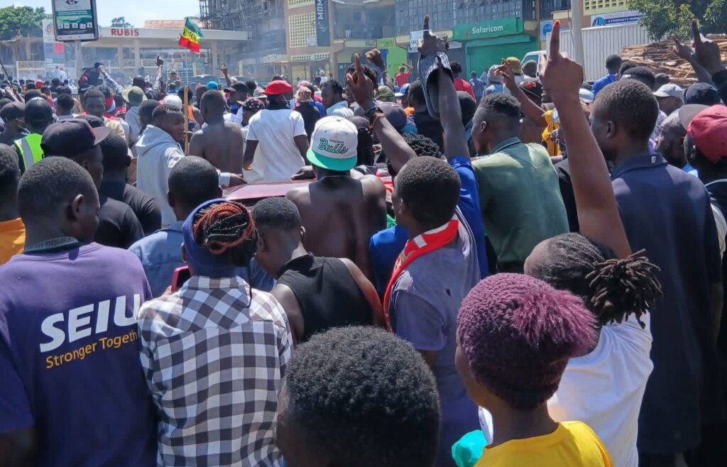 A large crowd of youths and residents gathers in Kakamega Town during a daytime protest, with many raising their hands and chanting along a busy street near shops and a petrol station.
