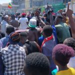 A large crowd of youths and residents gathers in Kakamega Town during a daytime protest, with many raising their hands and chanting along a busy street near shops and a petrol station.