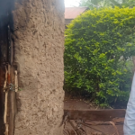 A partially burnt mud house with flames visible inside as smoke rises from the doorway, while a man stands nearby observing the damage in Mabanga, Bukembe West, Bungoma County.
