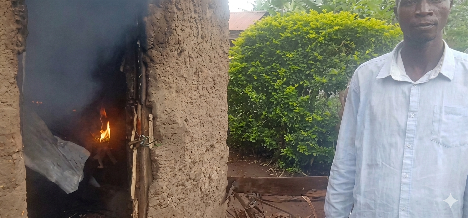 A partially burnt mud house with flames visible inside as smoke rises from the doorway, while a man stands nearby observing the damage in Mabanga, Bukembe West, Bungoma County.
