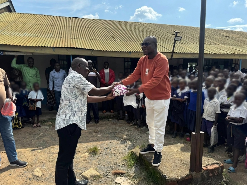 Philip Etale presents a football to a school official at Munyuki Comprehensive School while dozens of pupils gather around during the handover event.