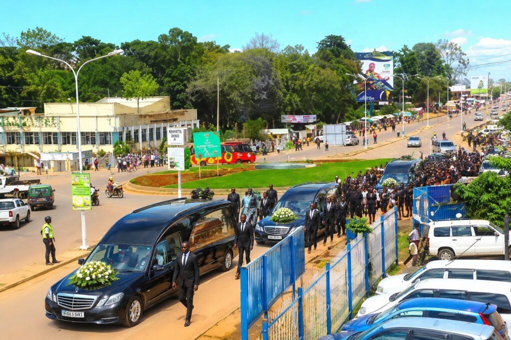 Several black hearses accompanied by a large group of mourners move along a busy road in Kakamega town as police officers and motorists look on near a roundabout and roadside buildings.