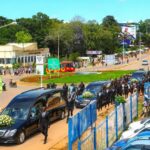 Several black hearses accompanied by a large group of mourners move along a busy road in Kakamega town as police officers and motorists look on near a roundabout and roadside buildings.