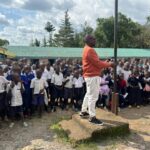 Philip Etale stands in front of a large group of pupils at Munyuki Comprehensive School while speaking during a school event.