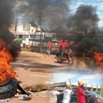 Protesters block a road in Mumias town using burning tyres and stones as thick black smoke rises into the sky while motorists and residents move through the tense scene.