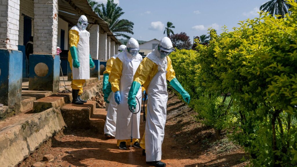 Four health workers dressed in protective hazmat suits, gloves, masks, and goggles walk along a path beside a building while carrying spraying equipment during an Ebola response or disease-control operation in a tropical setting.