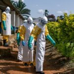 Four health workers dressed in protective hazmat suits, gloves, masks, and goggles walk along a path beside a building while carrying spraying equipment during an Ebola response or disease-control operation in a tropical setting.