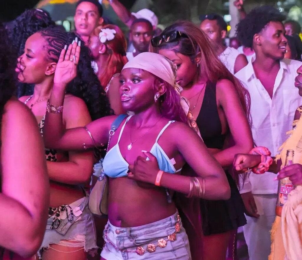 A crowd of young revellers at the Summer Tides Festival in Malindi dances under colorful stage lighting. Several attendees are dressed in beachwear and casual party outfits as they enjoy the music in a packed outdoor venue.