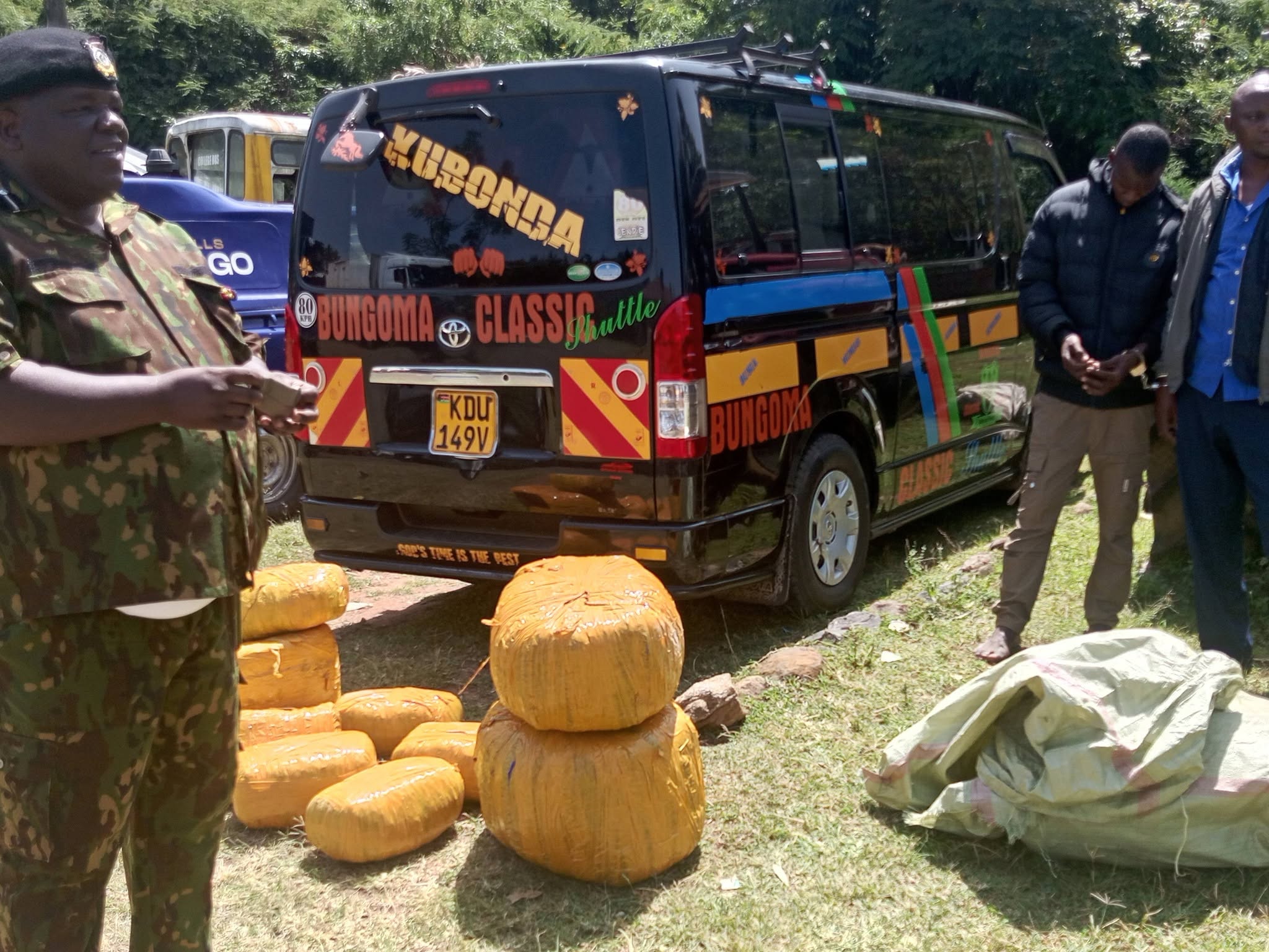Police officers stand beside a black public service van after intercepting it at Lumakanda Junction. Several yellow-wrapped bales of cannabis sativa and green sacks containing the drugs are displayed on the ground following a Sh3 million narcotics seizure.
