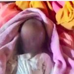 A three-week-old infant wrapped in a pink blanket, lying on a bed with colorful blankets in the background. The child's face is blurred to protect their identity.