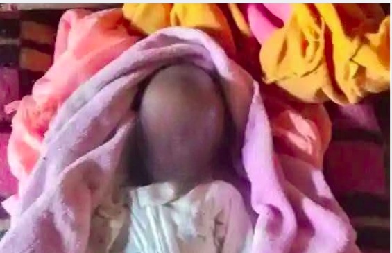 A three-week-old infant wrapped in a pink blanket, lying on a bed with colorful blankets in the background. The child's face is blurred to protect their identity.