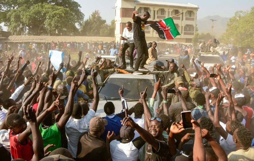 A large crowd of political supporters surrounds a vehicle in a dusty street, with several people standing on its roof and waving a Kenyan flag as others raise their hands and record the scene on mobile phones.