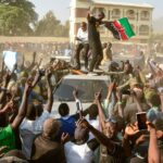 A large crowd of political supporters surrounds a vehicle in a dusty street, with several people standing on its roof and waving a Kenyan flag as others raise their hands and record the scene on mobile phones.