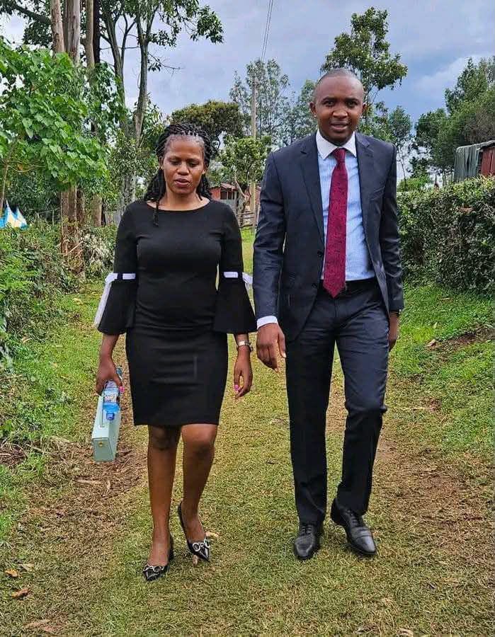 A man and woman walking together on a dirt path lined with greenery and eucalyptus trees. She wears a black bell-sleeve dress with white cuff trim, patterned pointed-toe heels, and a bracelet, and carries a light green box in one hand. He wears a dark suit with a light blue shirt and a maroon tie, paired with black dress shoes. They are holding hands, with hedges, trees, and a cloudy sky in the background.
